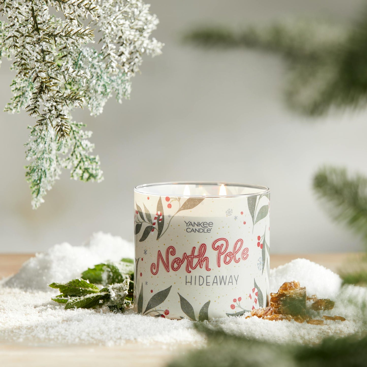 north pole hideaway three wick candle on surface with snow, green leaves, amber, and evergreen twigs