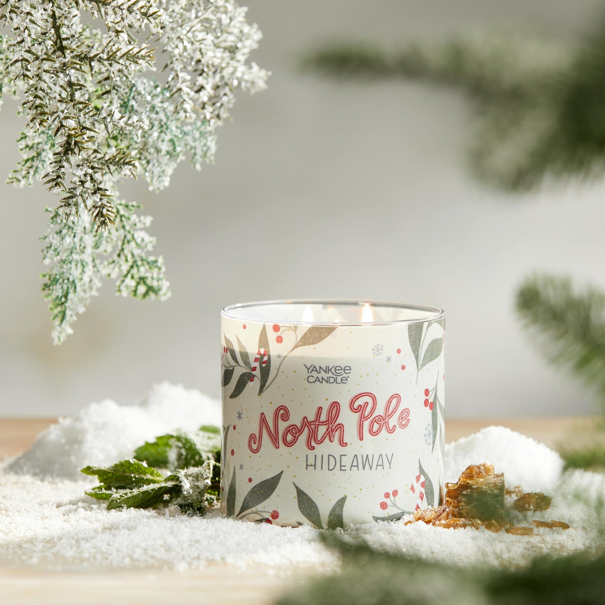north pole hideaway three wick candle on surface with snow, green leaves, amber, and evergreen twigs