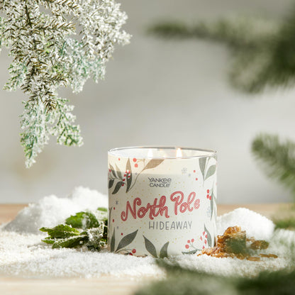 north pole hideaway three wick candle on surface with snow, green leaves, amber, and evergreen twigs