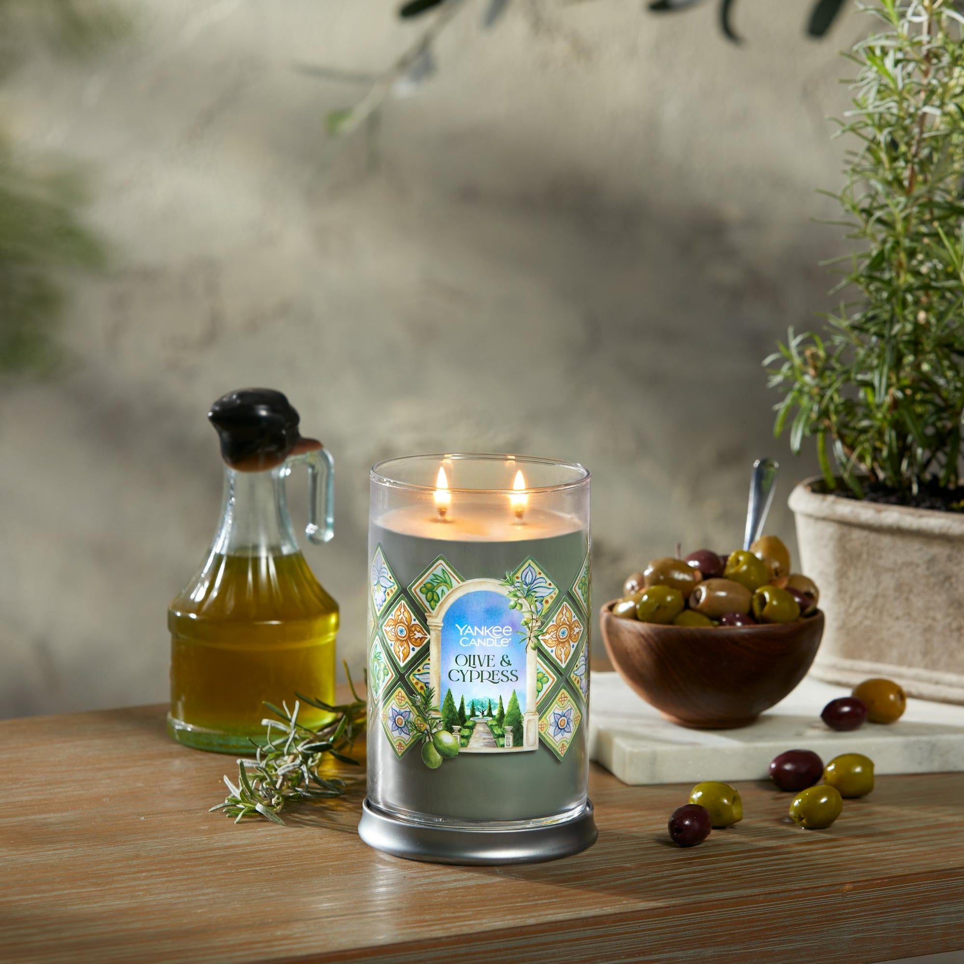 Hello, Italy Collection Olive & Cypress Signature Large Tumbler Candle on a table with a jar of olive oil and a bowl of olives