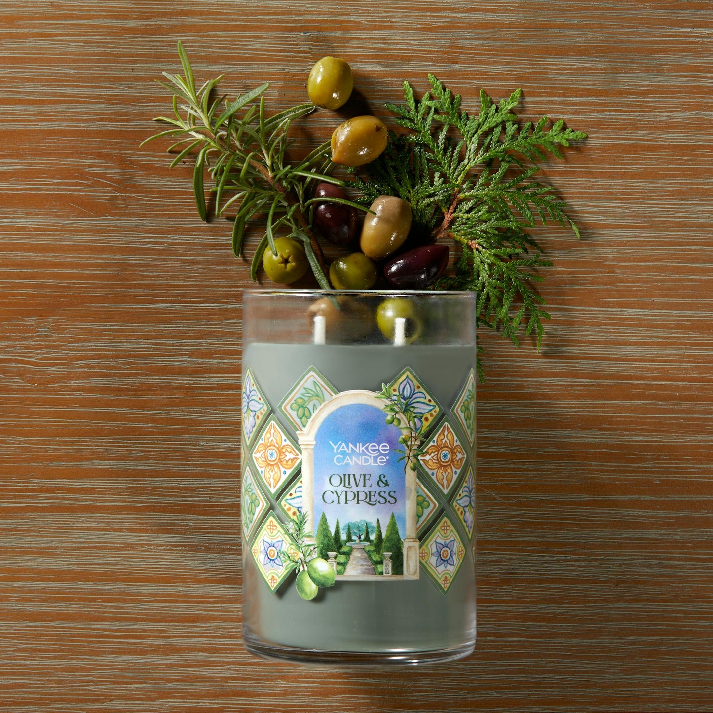 Hello, Italy! Collection Olive & Cypress Signature Large Tumbler Candle laying on a table with olives and sprigs of green leaves coming from the top of the candle