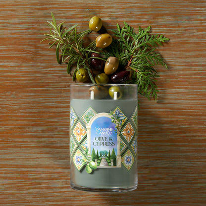 Hello, Italy! Collection Olive & Cypress Signature Large Tumbler Candle laying on a table with olives and sprigs of green leaves coming from the top of the candle