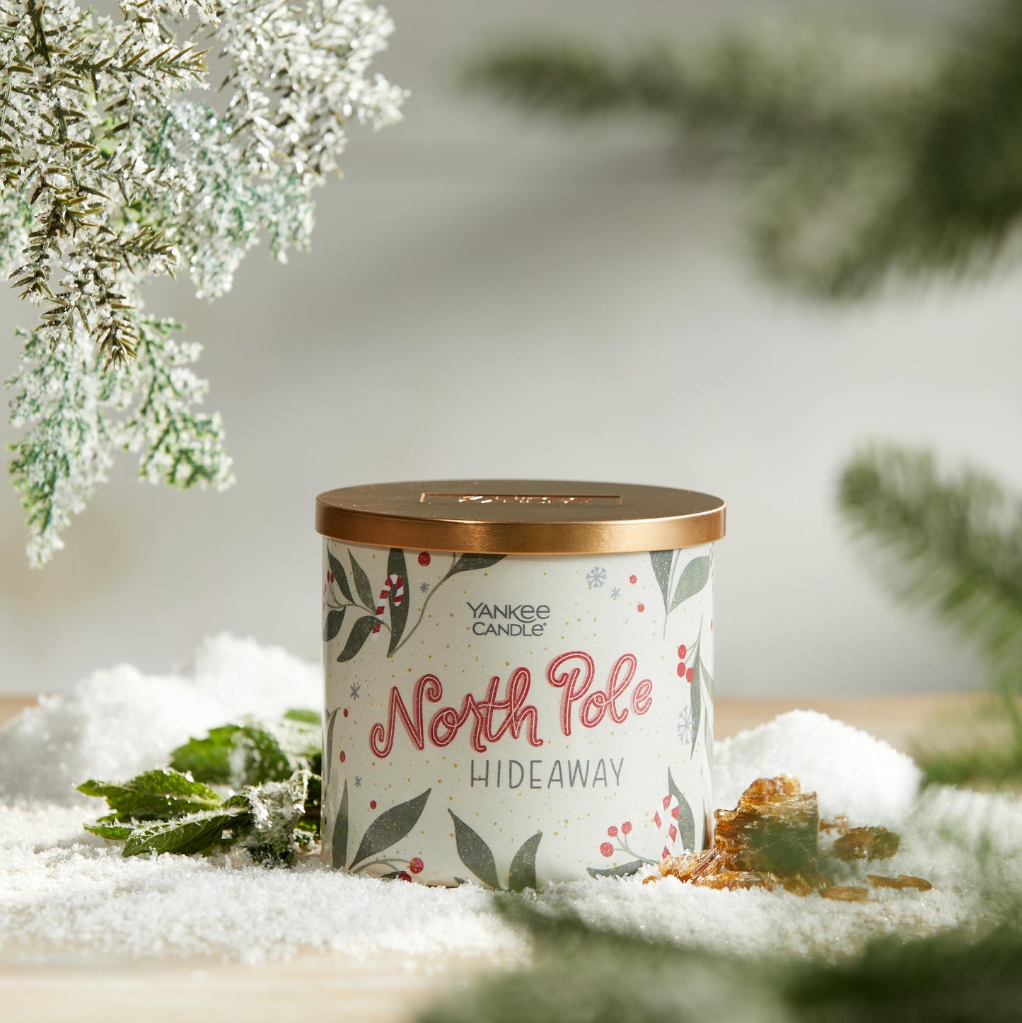 north pole hideaway three wick candle on surface with snow, green leaves, amber, and evergreen twigs