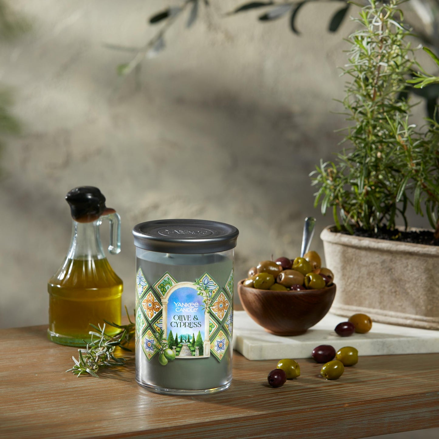 Hello, Italy! Collection Olive & Cypress Signature Large Tumbler Candle on a table with a jar of olive oil and a bowl of olives