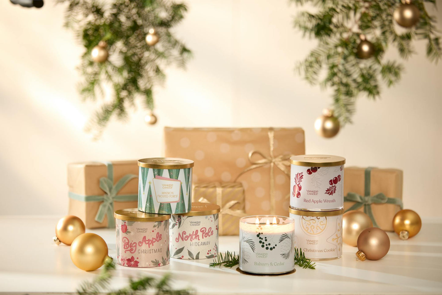 Magical Frosted Forest 3-Wick Candle, Balsam & Cedar 3-Wick Candle, Red Apple Wreath 3-Wick Candle, and Christmas Cookie 3-Wick Candle with Passport to the Holidays Collection Big Apple Christmas 3-Wick Candle and North Pole Hideaway 3-Wick Candle on a table next to wrapped gifts and gold ornaments