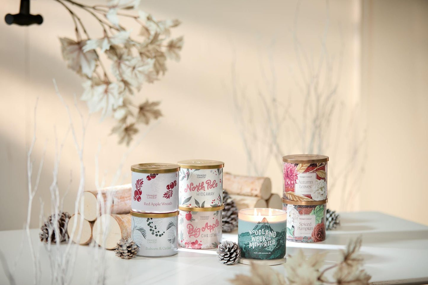 Red Apple Wreath 3-Wick Candle, Balsam & Cedar 3-Wick Candle, Autumn Leaves 3-Wick Candle, and Spiced Pumpkin 3-Wick Candle next to Passport to the Holidays Collection North Pole Hideaway 3-Wick Candle and Big Apple Christmas 3-Wick Candle next to a Cottage Getaway Collection Woodland Weekend Memories 3-Wick Candle on a table